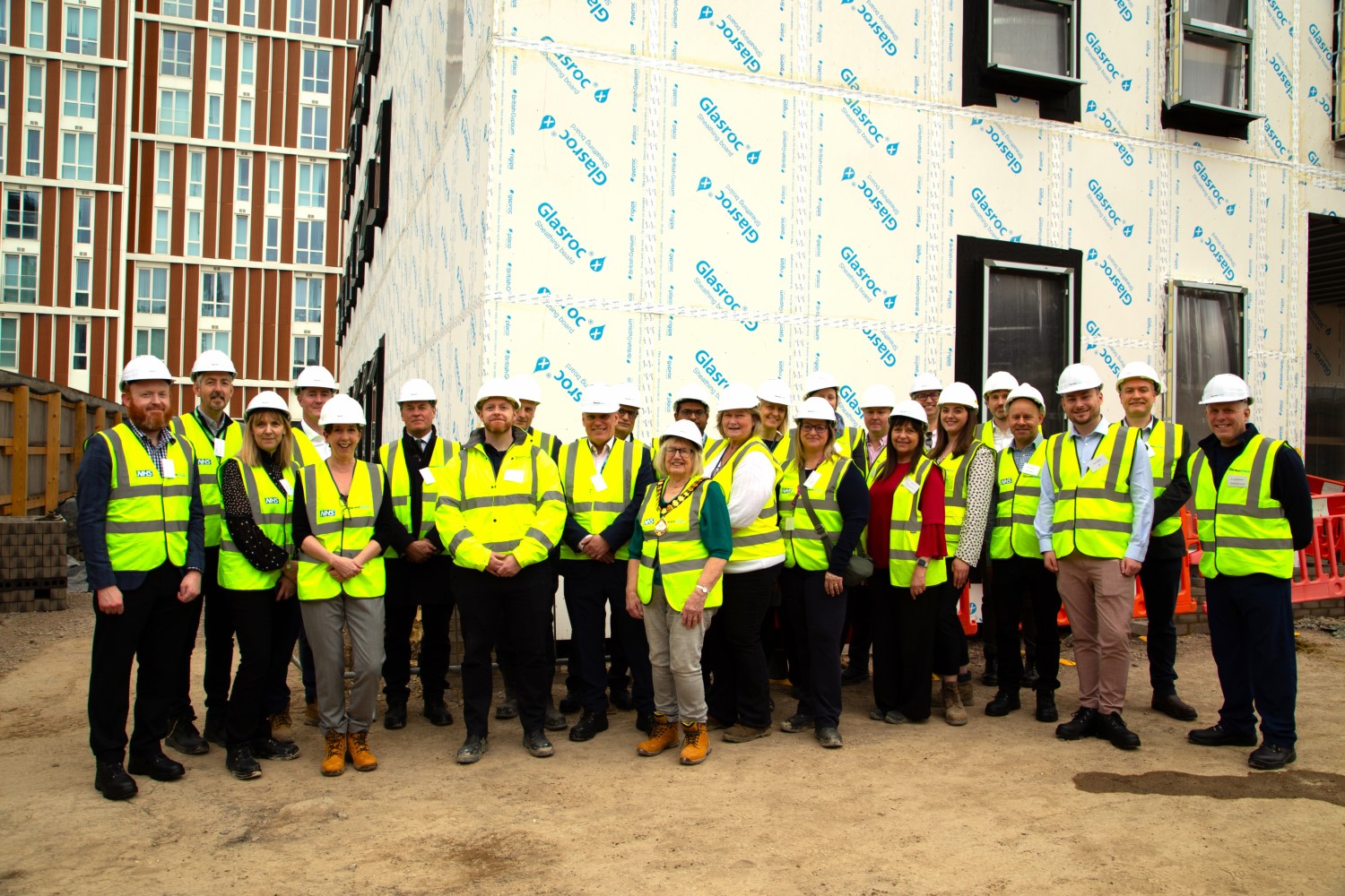A group of people of different genders and race are stood in front of an unfinished building. They are wearing high vis vests and hard hats.
