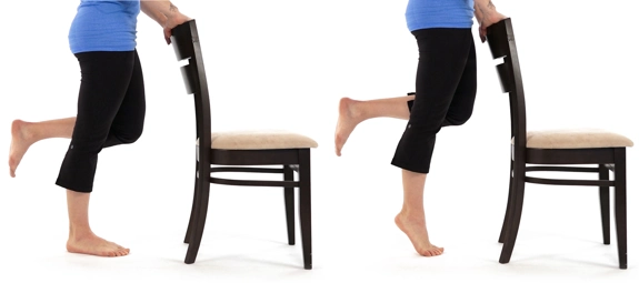 Person standing behind chair, holding the top, with one leg up, and rising to tiptoes on the other