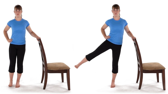 Person standing side-on to a chair, using the back for support while other hand is on hip, then raising the outer leg sideways.