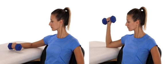 Person sitting side-on next to a table with elbow resting on it while holding a dumbbell. They lift the dumbbell at a right angle.