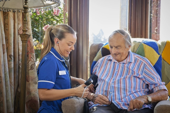 A community nurse taking a patient's blood pressure, while they sit in a comfy chair at home