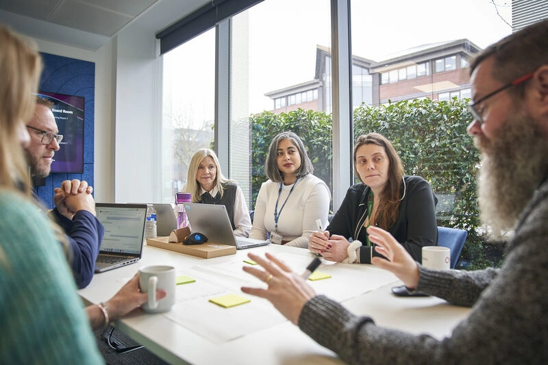 A group of 6 staff members, sitting around a table in a meeting