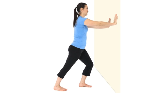 Person standing with split leg stance (one leg straight and the other bent in front) and pushing against wall