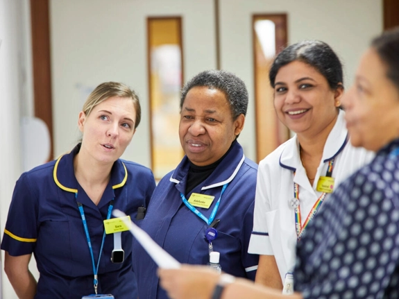 Four healthcare staff stand together in a hospital corridor, discussing something as one holds a sheet of paper.