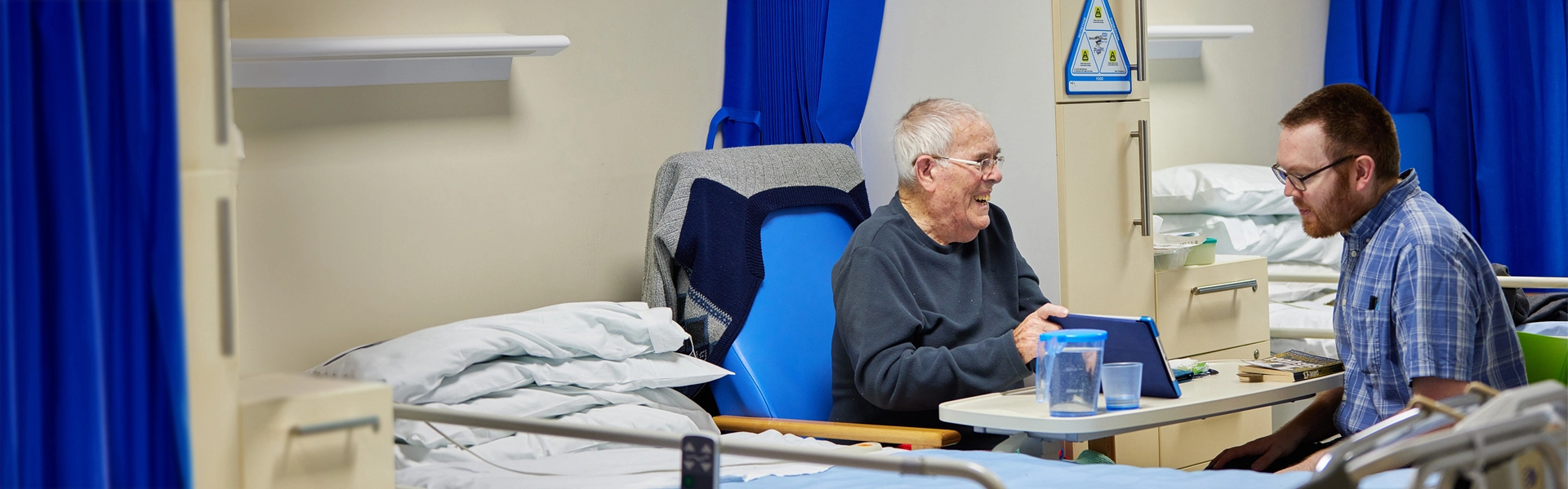 patient sitting by bed showing man something on his tablet