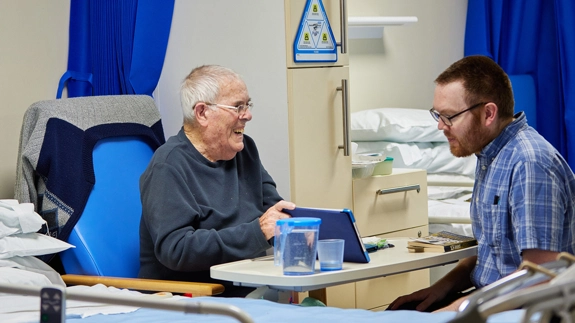 patient sitting in a ward chair, showing a staff member something on his tablet