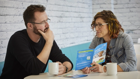 Two people discussing a leaflet while sitting at a table with a hot drink