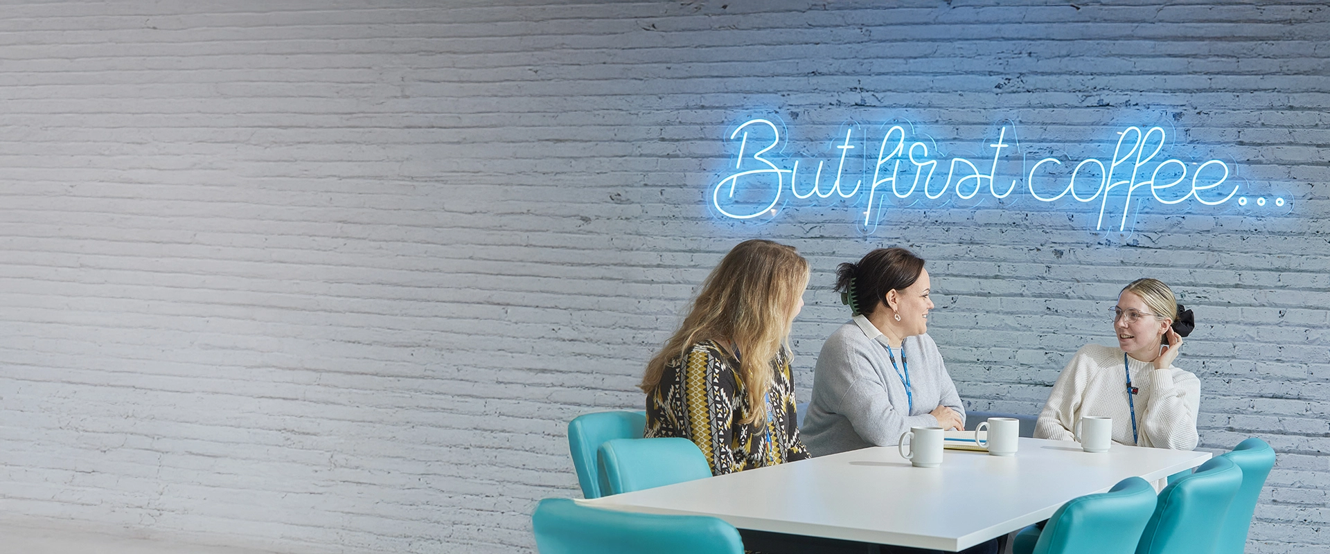 Three staff members, sat at a table under a neon sign that says 'But first coffee...'